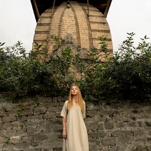 Puede incluir: Una mujer con un vestido largo de lino marr&oacute;n claro se encuentra frente a un muro de piedra con un gran edificio antiguo de ladrillo detr&aacute;s de ella. El edificio tiene un techo en forma de c&uacute;pula y tallas intrincadas.