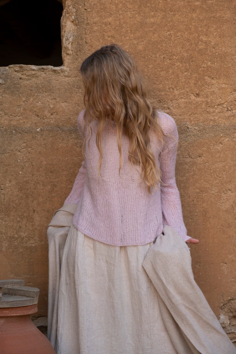 May include: A woman wearing a light pink knitted sweater and a long, flowing beige skirt. She is standing in front of a textured, tan wall.