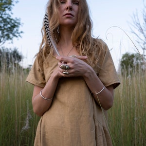 May include: A woman with long blonde hair wearing a brown linen dress and silver bracelets. She is holding a feather in her hand and has several rings on her fingers.
