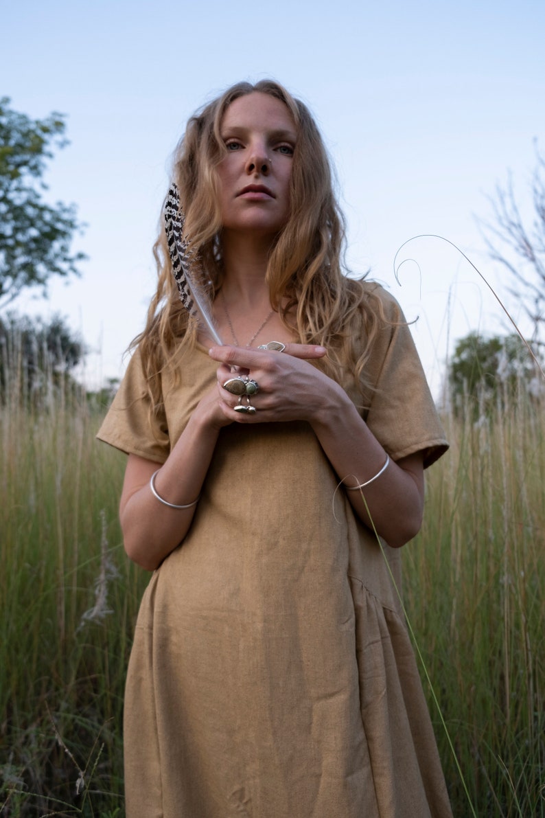 May include: A woman with long blonde hair wearing a brown linen dress and silver bracelets. She is holding a feather in her hand and has several rings on her fingers.