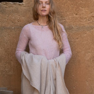 May include: A woman wearing a pink knitted sweater and a beige shawl. She is standing in front of a textured wall.