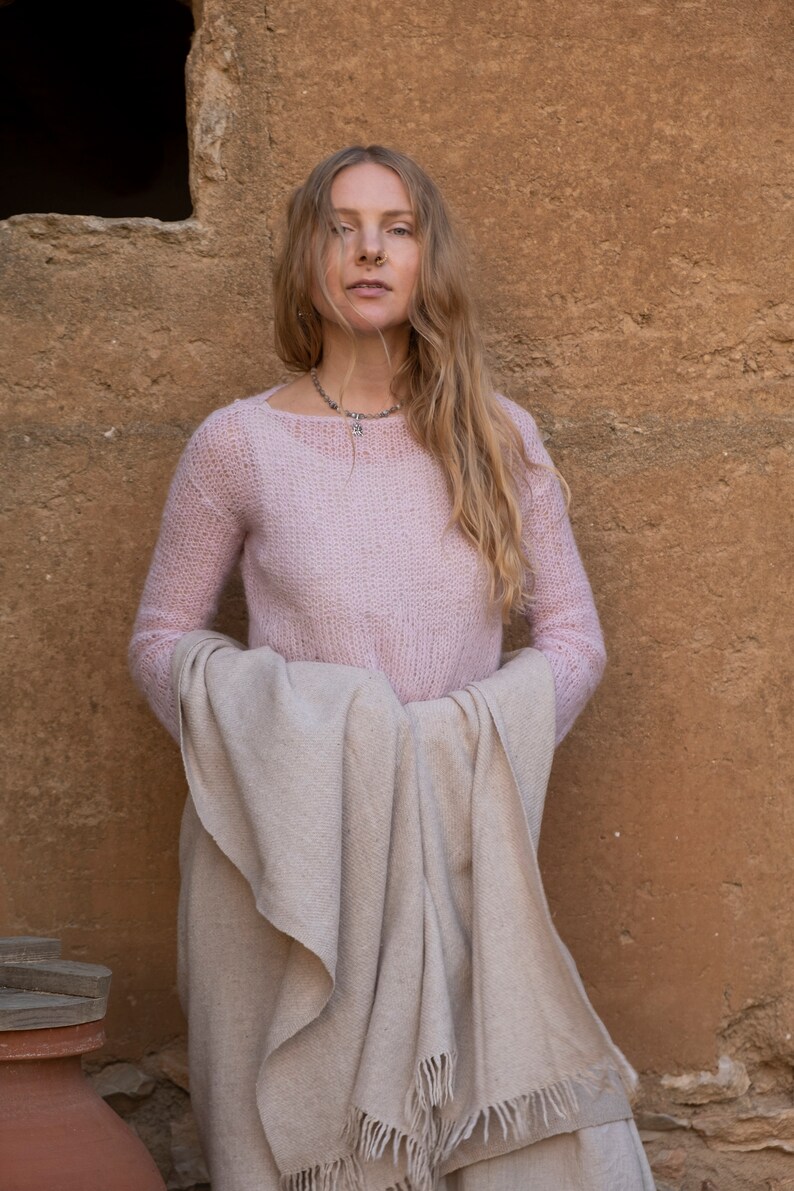 May include: A woman wearing a pink knitted sweater and a beige shawl. She is standing in front of a textured wall.