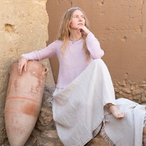May include: A woman wearing a light pink sweater and a long, flowing white skirt sits on a rock with a large clay pot beside her. She has long blonde hair and is looking off to the side.