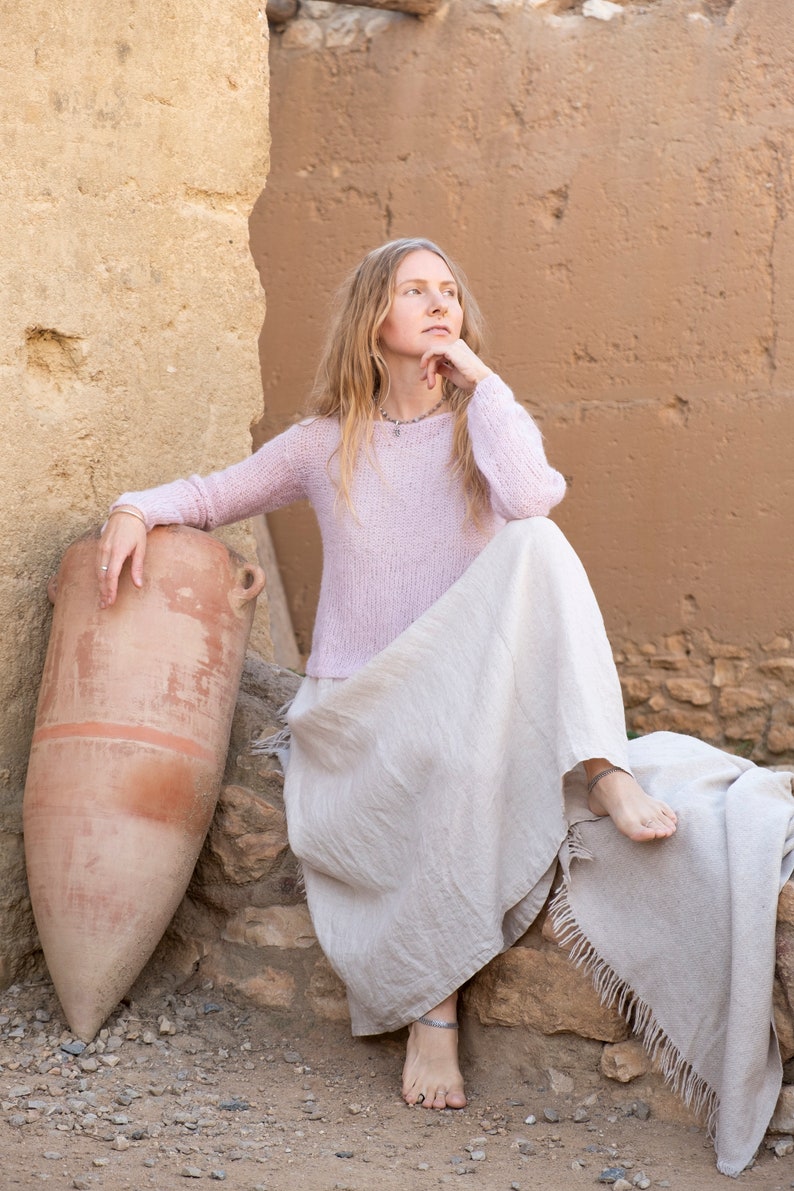 May include: A woman wearing a light pink sweater and a long, flowing white skirt sits on a rock with a large clay pot beside her. She has long blonde hair and is looking off to the side.