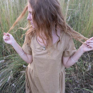 May include: A woman with long, curly, light brown hair wearing a light brown, short-sleeved dress with a silver necklace. She is standing in a field of tall grass.