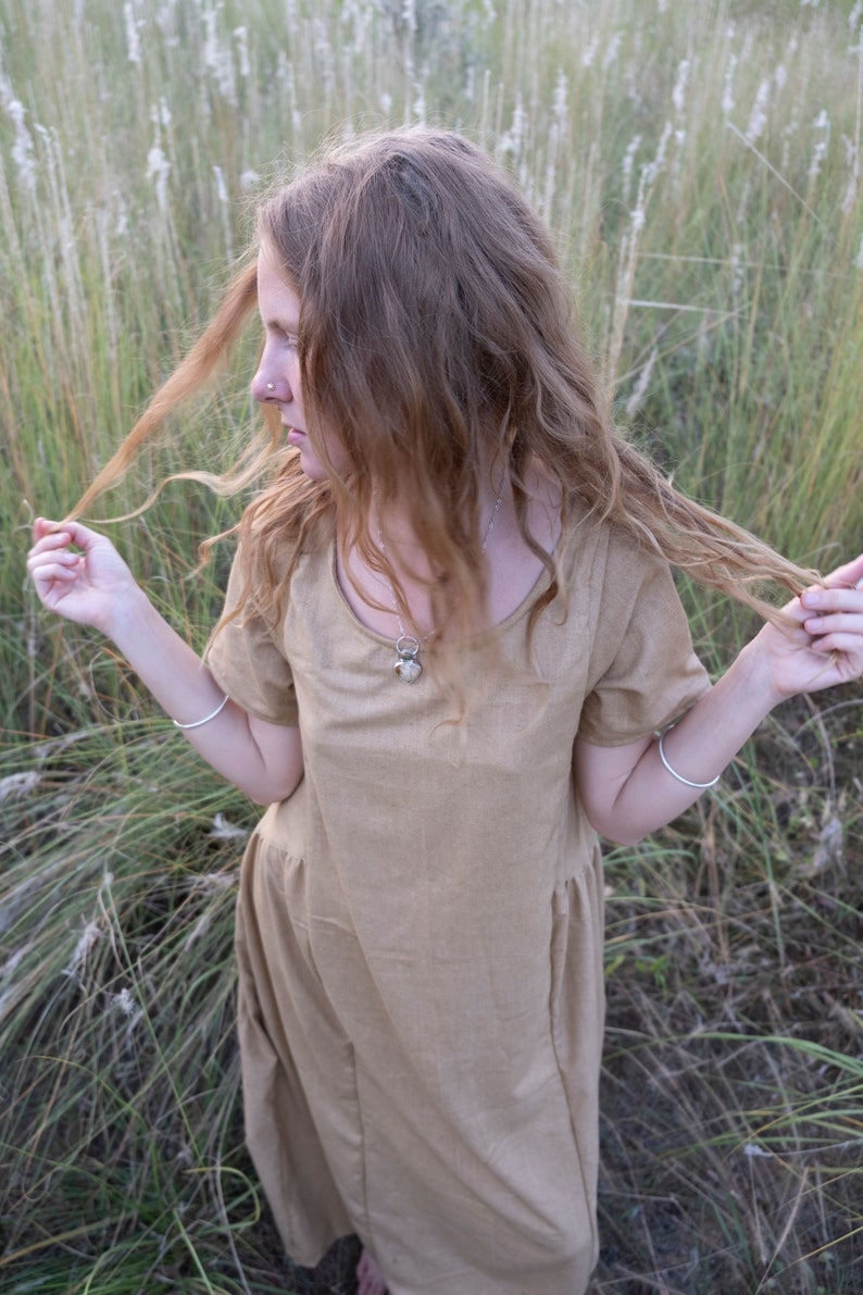 May include: A woman with long, curly, light brown hair wearing a light brown, short-sleeved dress with a silver necklace. She is standing in a field of tall grass.