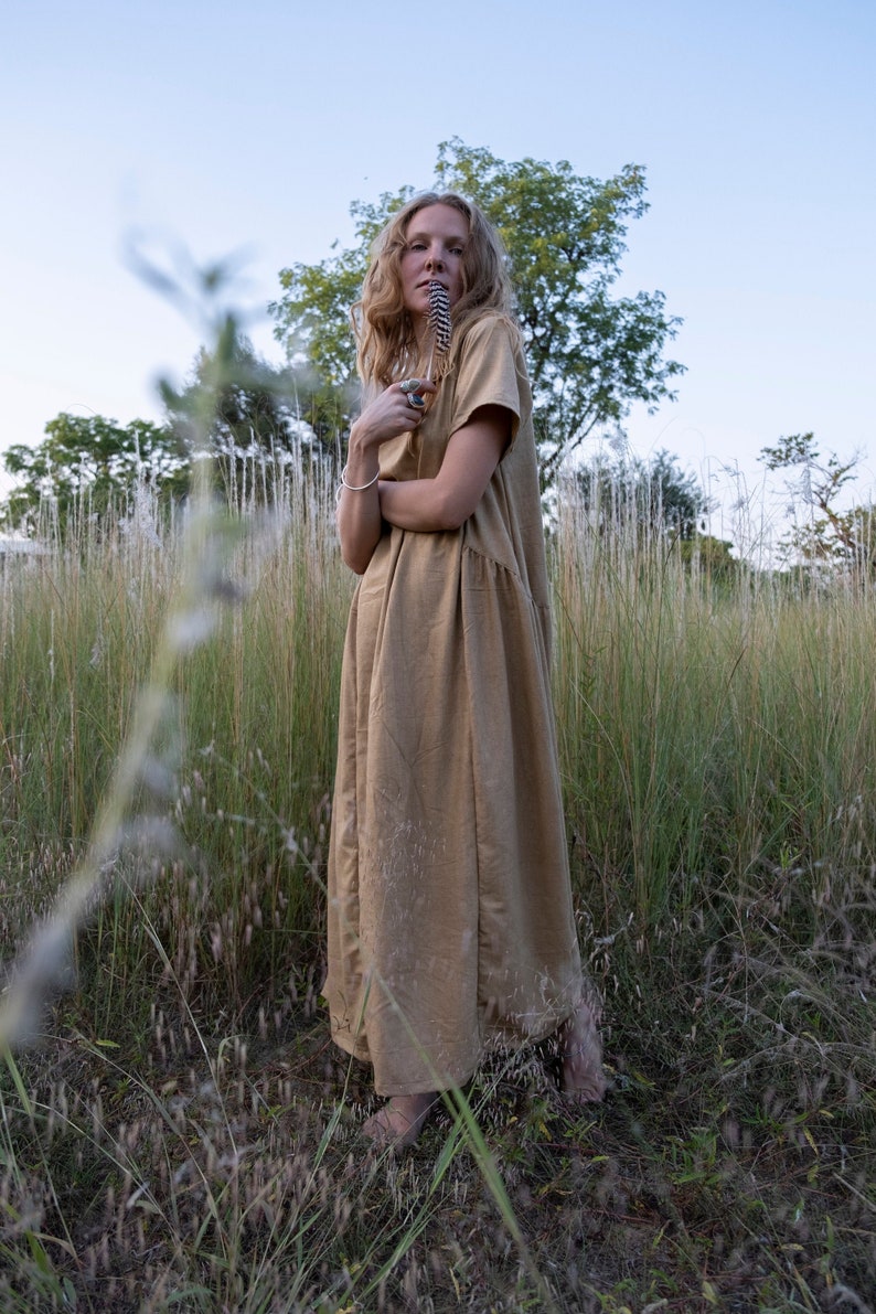 May include: A woman in a long, light brown dress stands in a field of tall grass. She is holding a feather in her mouth and looking up at the sky.