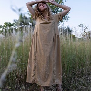 May include: A woman wearing a long, light brown linen dress stands in a field of tall grass. She is barefoot and has her arms raised above her head.