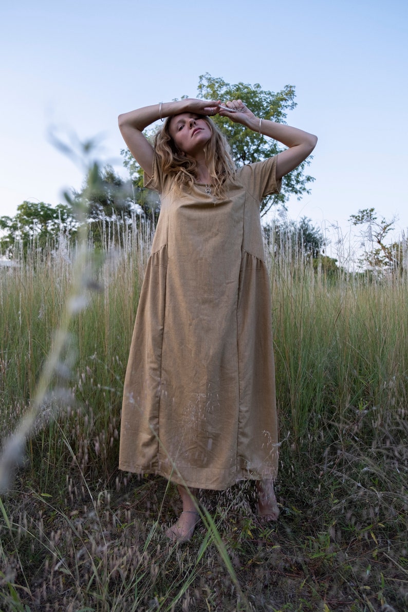 May include: A woman wearing a long, light brown linen dress stands in a field of tall grass. She is barefoot and has her arms raised above her head.