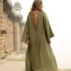 Puede incluir: Vestido largo maxi verde oliva con mangas largas y acampanadas y espalda baja. El vestido se usa al aire libre, con un edificio histórico al fondo. Una bufanda floral rosa se usa alrededor del cuello.