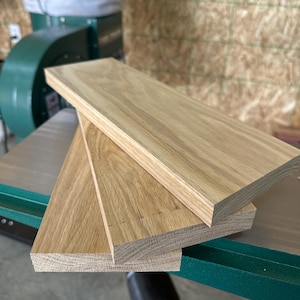 May include: Three unfinished, light-colored wooden planks are stacked on a green surface. The planks have a natural wood grain and are rectangular in shape. The image suggests a woodworking project or the sale of lumber.