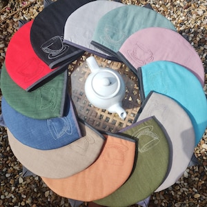 Op de afbeelding: Een set van 10 kleurrijke theekopwarmers met geborduurde theekopjesontwerpen. De warmers zijn gemaakt van stof en zijn in verschillende kleuren, waaronder blauw, groen, roze en bruin. De warmers zijn in een cirkel rond een witte theepot gerangschikt.