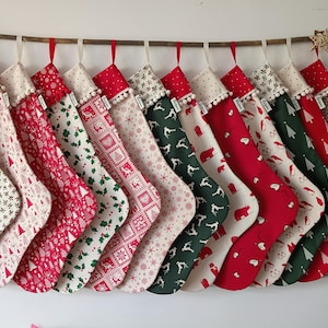 Könnte beinhalten: Eine Reihe von zwölf Weihnachtsstrümpfen, die an einem Ast hängen. Jeder Strumpf hat ein einzigartiges festliches Muster in Rot, Grün und Creme, mit Motiven von Bäumen, Bären und Schneeflocken, verziert mit einer cremefarbenen Bommelborte.
