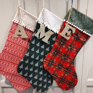 May include: Three Christmas stockings in red, green and white fabric. The stockings are decorated with a plaid pattern, a Christmas tree pattern and white polka dots. Each stocking has a wooden letter attached to it, spelling out the word "AME".