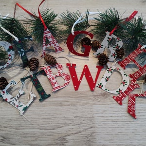 May include: Festive wooden letter ornaments with Christmas patterns and red ribbons. The letters, spelling "FAB WORE", are displayed with pinecones and evergreen branches on a light wood surface. Each letter has a unique design.