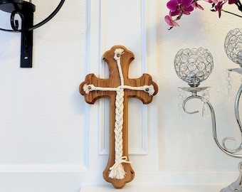 May include: A wooden cross with a white braided rope design. The cross is mounted on a white wall and has a natural wood finish.