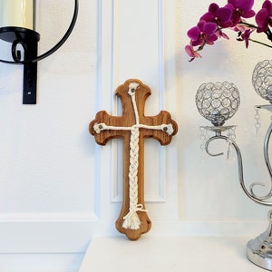 May include: A wooden cross with a white braided rope design. The cross is mounted on a white wall and has a natural wood finish.