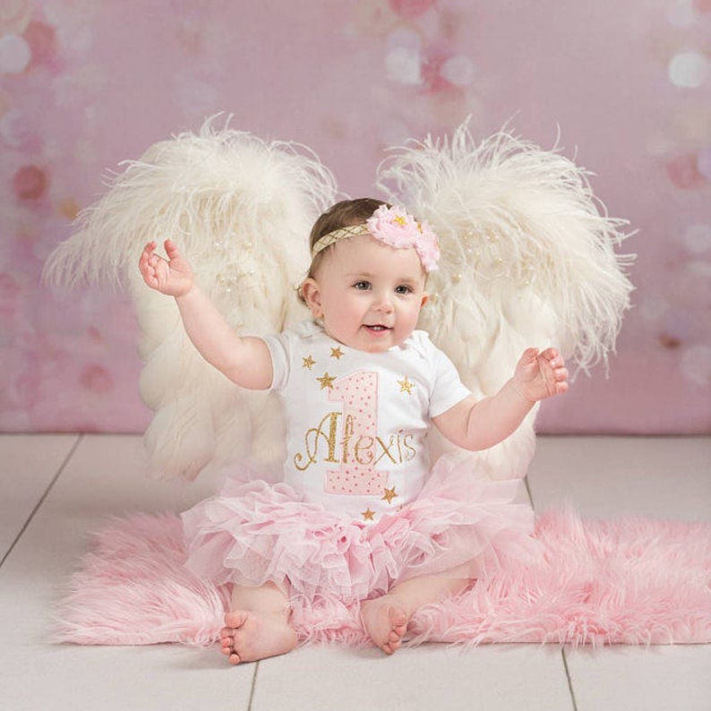 Puede incluir: Una ni&ntilde;a peque&ntilde;a con un body blanco con estrellas doradas brillantes y el n&uacute;mero uno con el nombre Alexis. Lleva una falda de tul rosa y una diadema de &aacute;ngel con alas de plumas blancas. Est&aacute; sentada en una alfombra peluda rosa.