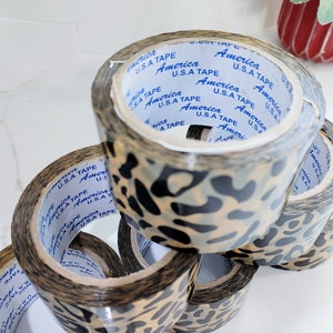 Puede incluir: Un rollo de cinta de embalaje de estampado de leopardo. Cada rollo tiene el texto "America U.S.A Tape" impreso en &eacute;l.