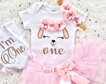 May include: A pink and white baby girl's first birthday outfit with a white onesie featuring a dog face and the text "one" in gold glitter, a white tutu skirt with a pink satin bow, a white diaper cover with the text "I'm One" in gold glitter, and a pink flower headband.