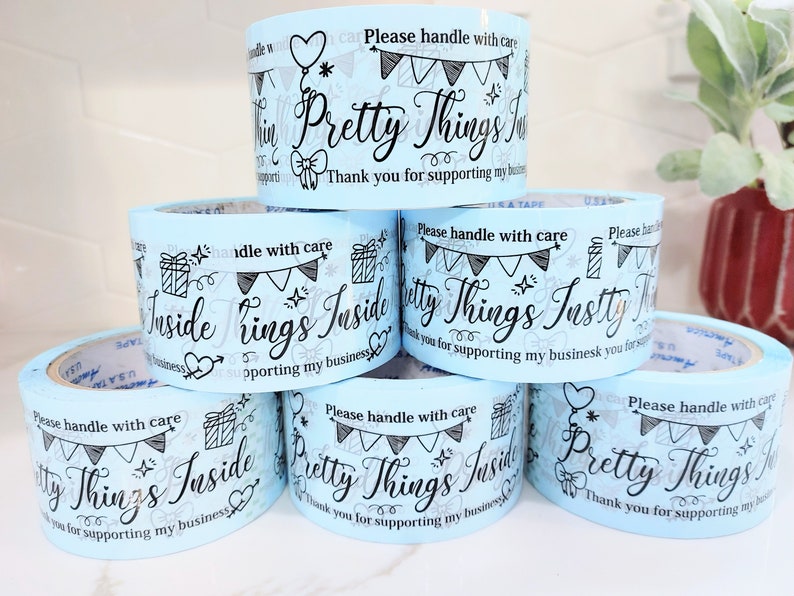 Puede incluir: Rollos de cinta de embalaje azul con el texto "Pretty Things Inside" y "Please handle with care" impreso en ellos. La cinta est&aacute; decorada con estrellas, corazones y banderas.