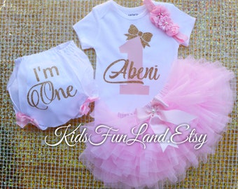 Puede incluir: Un conjunto de ropa de bebé blanco con una falda tutú rosa, diadema y cubrepañales. El conjunto incluye un body de manga corta con un lazo de purpurina dorada y el nombre "Abeni". El cubrepañales dice "I'm One".