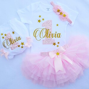 Puede incluir: Un conjunto de primer cumplea&ntilde;os rosa y dorado para una ni&ntilde;a. El conjunto incluye un body blanco con un n&uacute;mero uno rosa y dorado y el nombre "Olivia" en purpurina dorada, un pa&ntilde;al blanco con un n&uacute;mero uno rosa y dorado y el nombre "Olivia" en purpurina dorada, una falda de tul rosa con un lazo de estrella dorada y una diadema rosa con estrellas doradas.