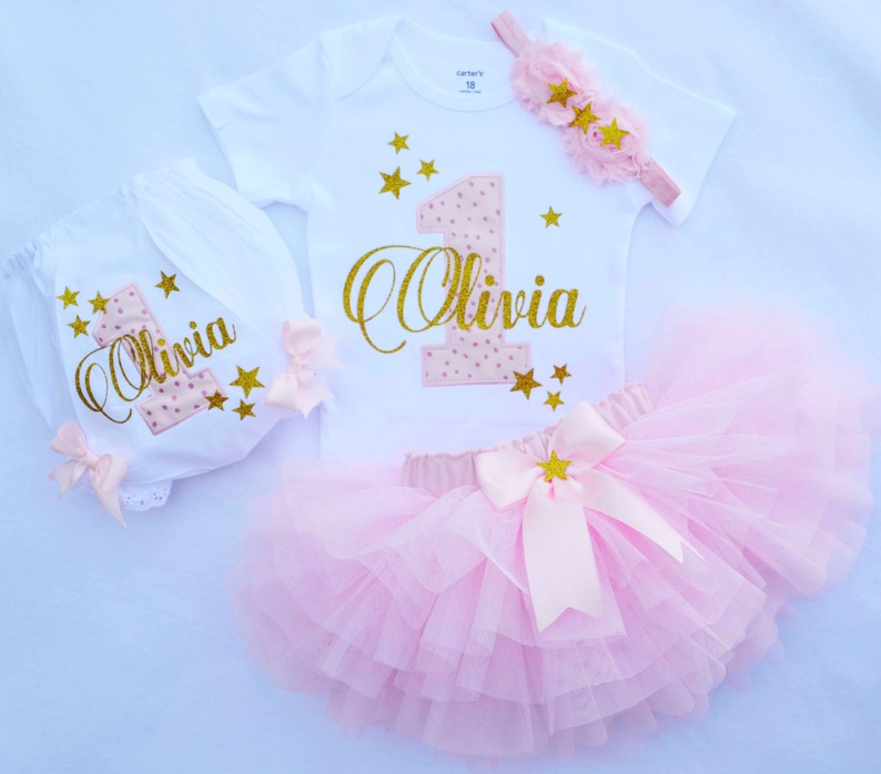Puede incluir: Un conjunto de primer cumplea&ntilde;os rosa y dorado para una ni&ntilde;a. El conjunto incluye un body blanco con un n&uacute;mero uno rosa y dorado y el nombre "Olivia" en purpurina dorada, un pa&ntilde;al blanco con un n&uacute;mero uno rosa y dorado y el nombre "Olivia" en purpurina dorada, una falda de tul rosa con un lazo de estrella dorada y una diadema rosa con estrellas doradas.