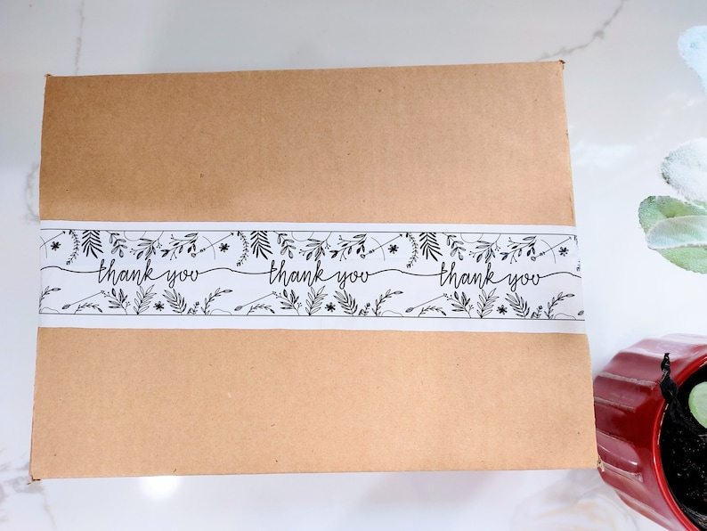 Puede incluir: Una caja de cart&oacute;n marr&oacute;n con una cinta de patr&oacute;n floral blanco y negro que dice "gracias" tres veces.