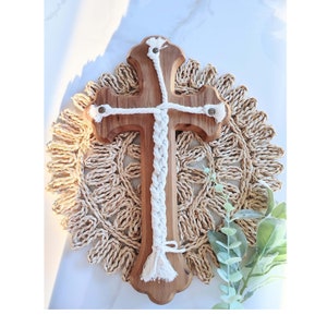 May include: A wooden cross with a white braided rope design. The cross is on a woven straw mat with green foliage.
