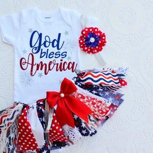 May include: A white baby onesie with the text "God bless America" in red and blue lettering with silver glitter accents and fireworks. A red, white, and blue fabric tutu with a red bow and a red, white, and blue flower headband.