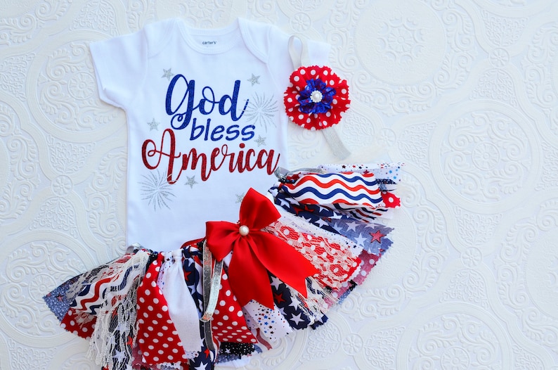 May include: A white baby onesie with the text "God bless America" in red and blue lettering with silver glitter accents and fireworks. A red, white, and blue fabric tutu with a red bow and a red, white, and blue flower headband.