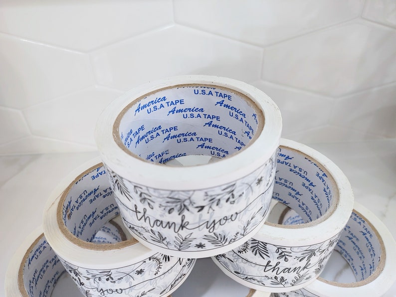 Puede incluir: Cuatro rollos de cinta de embalaje blanca con dise&ntilde;os florales en blanco y negro y el texto "gracias". La cinta est&aacute; etiquetada como "America U.S.A Tape".