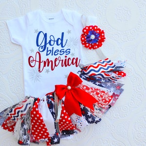 May include: A white baby onesie with the text "God bless America" in red and blue glitter. The onesie is paired with a red, white, and blue tutu with a red bow and a red, white, and blue flower headband.
