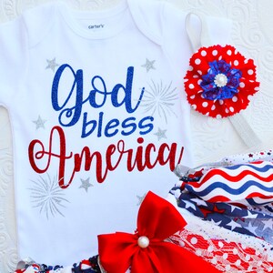 May include: A white baby onesie with the text "God bless America" in red, blue, and silver glitter. The onesie is paired with a red, white, and blue tutu and a headband with a red and white polka dot flower.