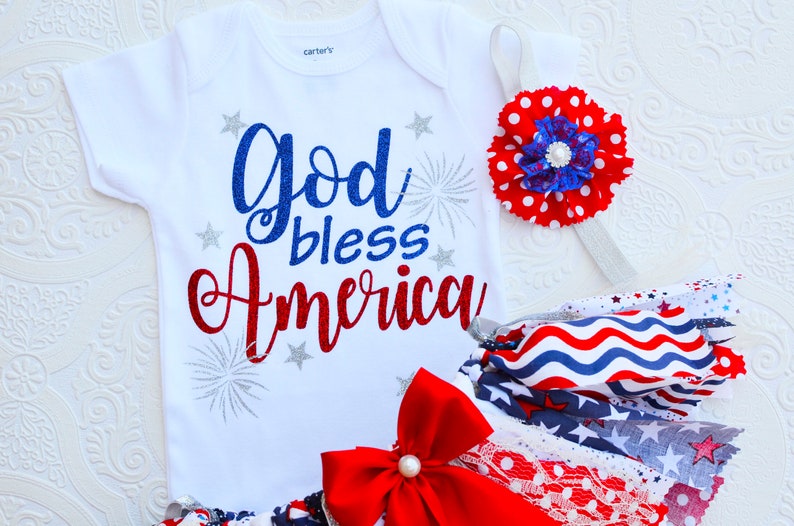 May include: A white baby onesie with the text "God bless America" in red, blue, and silver glitter. The onesie is paired with a red, white, and blue tutu and a headband with a red and white polka dot flower.
