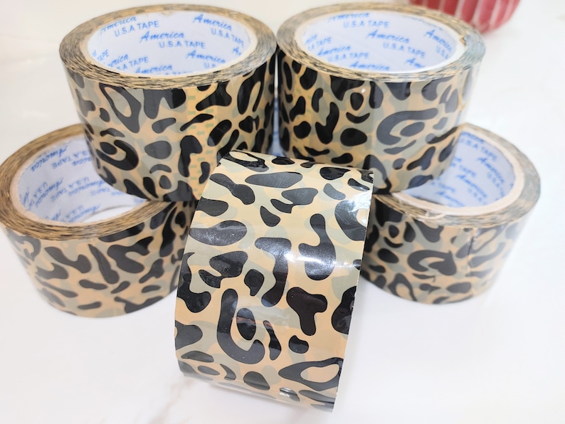 Puede incluir: Cuatro rollos de cinta adhesiva de estampado de leopardo. La cinta es marr&oacute;n y negra con un acabado brillante. La cinta est&aacute; etiquetada con "USA TAPE" y "America".