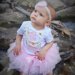Puede incluir: Una ni&ntilde;a peque&ntilde;a con un body blanco con un n&uacute;mero uno rosa y estrellas doradas brillantes. El body dice "Uno" y tiene lunares rosas. Tambi&eacute;n lleva una falda de tul rosa y un collar de perlas. Lleva una diadema de flores rosas en la cabeza.