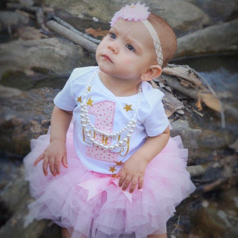 Puede incluir: Una ni&ntilde;a peque&ntilde;a con un body blanco con un n&uacute;mero uno rosa y estrellas doradas brillantes. El body dice "Uno" y tiene lunares rosas. Tambi&eacute;n lleva una falda de tul rosa y un collar de perlas. Lleva una diadema de flores rosas en la cabeza.
