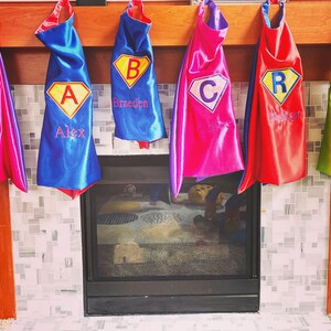 Puede incluir: Cinco capas de superhéroe colgadas en una repisa de chimenea. Las capas son rojas, azules, rosas, verdes y moradas. Cada capa tiene un rombo amarillo con una letra bordada en el centro. Las letras son K, A, C, R y L. Las capas están personalizadas con los nombres: Katie, Alex, Braeden, Ryker y L.