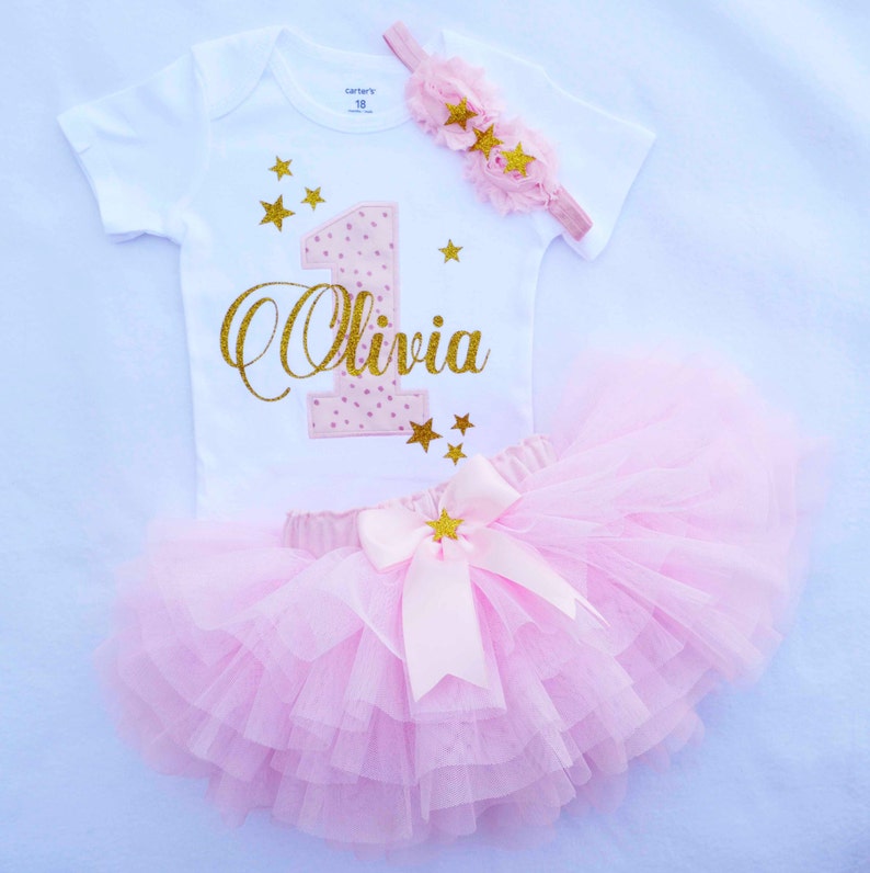 Puede incluir: Una falda de tul rosa con un lazo de sat&eacute;n rosa y estrellas doradas con purpurina. Un body blanco con un n&uacute;mero uno rosa y dorado con purpurina y el nombre "Olivia" en purpurina dorada. Una diadema rosa con estrellas doradas con purpurina.