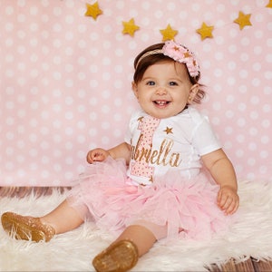 Puede incluir: Una ni&ntilde;a peque&ntilde;a con un body blanco con un n&uacute;mero uno rosa y dorado y el nombre "Gabriella" en purpurina dorada. Lleva una falda de tul rosa y una diadema de purpurina dorada con una flor rosa.