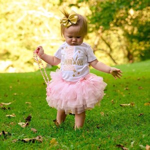 Puede incluir: Una ni&ntilde;a peque&ntilde;a con una camiseta blanca con purpurina dorada y el texto "Anniston" en ella. Tambi&eacute;n lleva una falda de tul rosa y un lazo dorado en el pelo. Est&aacute; sosteniendo un collar de cuentas rosas.