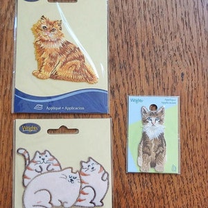 May include: Three cat-themed iron-on appliques on cardboard backing. One applique features a fluffy orange cat, another shows three cats, and the third depicts a small cat. The brand "Wrights" is printed on the top of each package.