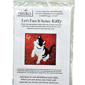 May include: A clear plastic package containing a fabric portrait kit. The kit features a black and white cat with a blue collar on a red background. Text on the package reads "Let's Face It Series: Kitty" and "Finished size 12"x14".