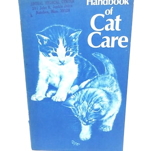 May include: A blue handbook titled "Handbook of Cat Care" featuring illustrations of two kittens. The cover also includes the text "ANIMAL MEDICAL CENTER" and an address. The kittens are white and gray.