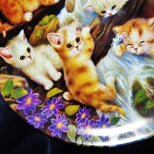 May include: A decorative plate featuring a colorful illustration of playful kittens. The kittens are depicted in shades of orange, white, and brown, amidst a backdrop of flowers and foliage. The plate has a glossy finish.