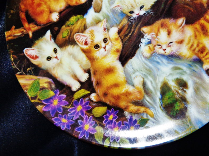 May include: A decorative plate featuring a colorful illustration of playful kittens. The kittens are depicted in shades of orange, white, and brown, amidst a backdrop of flowers and foliage. The plate has a glossy finish.
