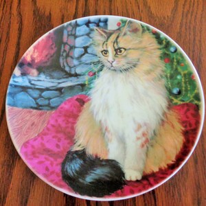 May include: A decorative plate featuring a fluffy cat with orange, white, and black fur. The cat is seated on a pink cushion in front of a fireplace and a Christmas tree. The plate has a white rim.