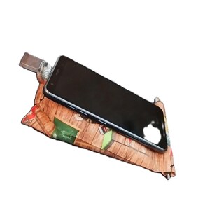 May include: A black smartphone rests on a patterned pillow-like phone stand. The stand features a brown, wood-grain design with green and red accents. The phone stand is designed to prop up a phone for hands-free use.